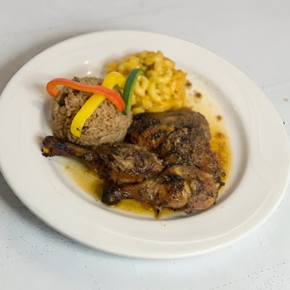 A plate of Jamaican jerk chicken with rice and peas, topped with bell peppers, and a side of macaroni and cheese.