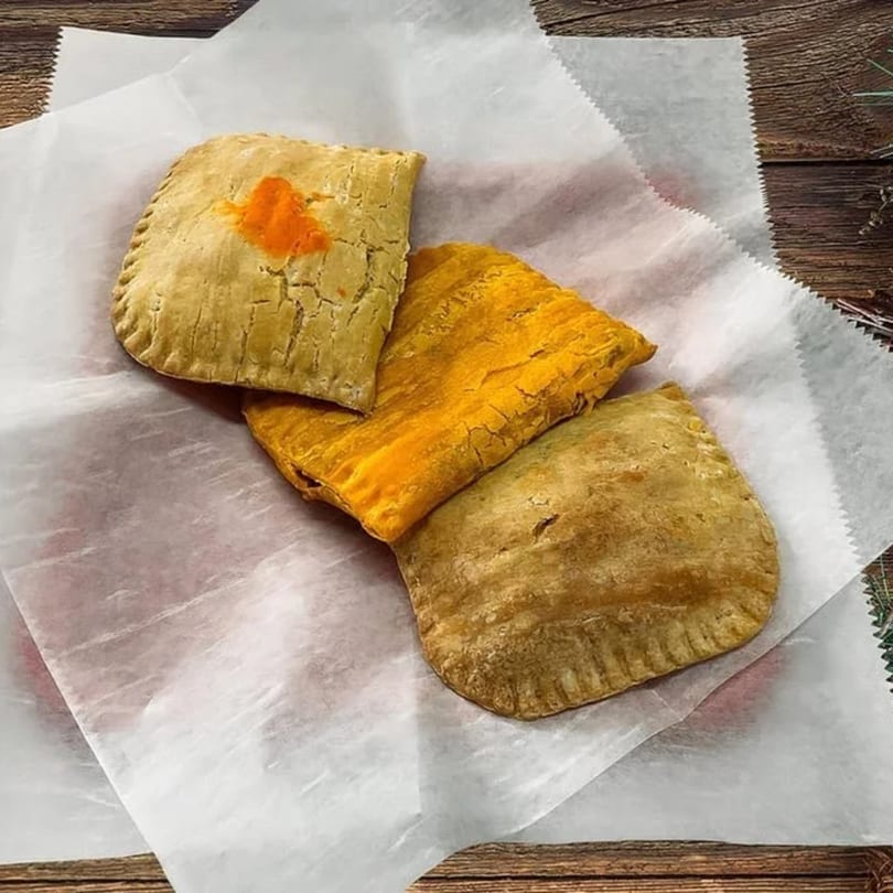 Three golden Jamaican patties, one with an orange spot, on white parchment paper over a wooden surface.